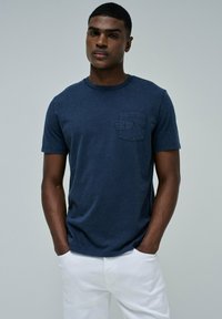 Camiseta de manga corta en azul marino con cuello redondo y un bolsillo en el pecho que presenta texto en relieve. Hecha de tela suave, con un corte relajado.