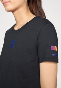 Musta puuvillapaita, jossa on sininen FC Barcelonan logo rinnassa ja värikkäällä raidallisella tikkauksella, jossa on Nike-logo hihassa.