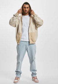 Beige zip-up hoodie med en stor hætte, frontlommer og ribbede manchetter, parret med lyseblå jeans og brune sneakers.