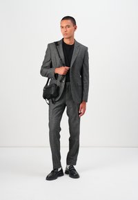 Grau maßgeschneiderter Anzug aus strukturiertem Stoff, schmaler Schnitt, kombiniert mit einem schwarzen Hemd. Zubehör: kleine schwarze Tasche und schwarze Schuhe.