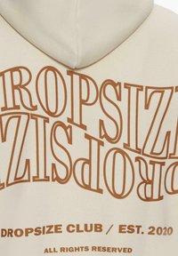Cream hoodie featuring large, upside-down brown text that reads "DROPSIZE CLUB / EST. 2020" with a smooth texture and a front pocket.