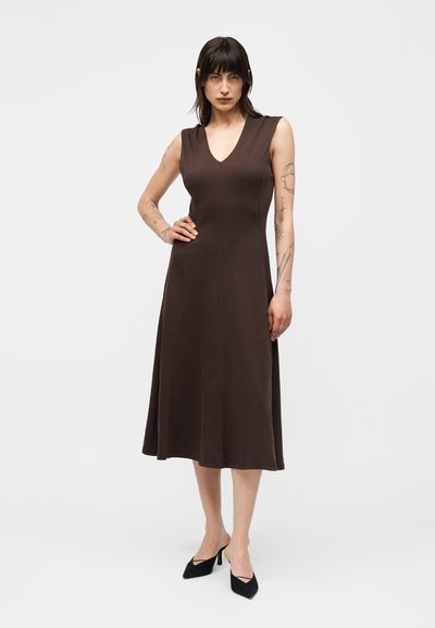 Γυναίκα με σκούρα μαλλιά που φοράει μαν sleeveless καφέ midi φόρεμα και μαύρα μυτερά τακούνια, στέκεται με ένα χέρι στη μέση σε απλό φόντο.