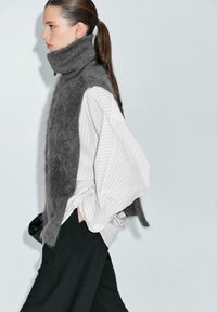 Gilet grigio peloso sopra una blusa bianca a righe, abbinato a pantaloni neri. Il gilet presenta un colletto alto e una vestibilità morbida.