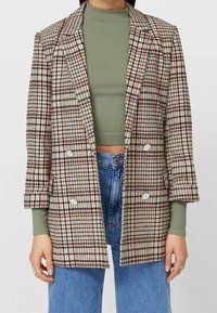 Blazer doppiopetto scozzese in beige, marrone e rosso. Presenta revers a punta, due grandi bottoni e una vestibilità ampia, abbinato a un top verde.