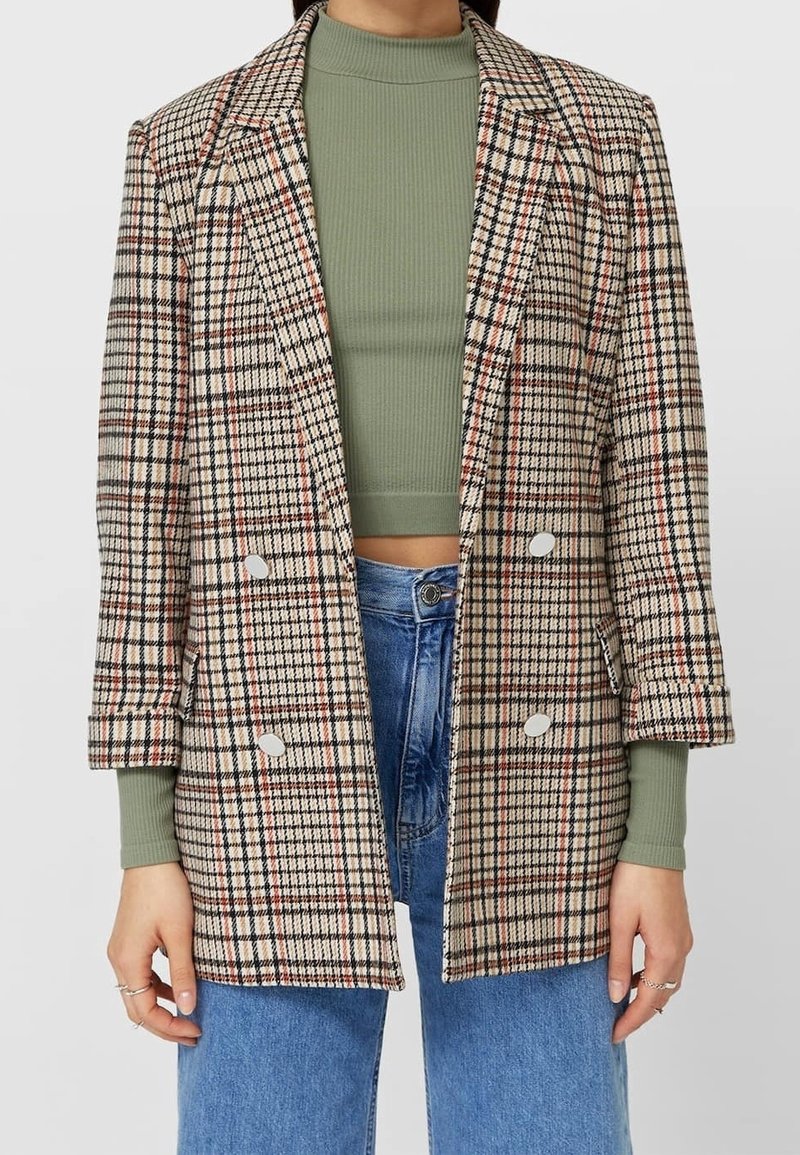 Blazer doppiopetto scozzese in beige, marrone e rosso. Presenta revers a punta, due grandi bottoni e una vestibilità ampia, abbinato a un top verde.