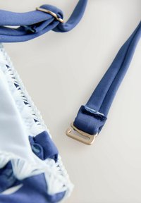 Nahaufnahme eines blauen verstellbaren BH-Trägers mit goldfarbenem Metallhaken und weißem Spitzenstoff vor hellem Hintergrund.