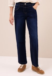 Persona vestida con jeans de pierna recta de color azul oscuro, camisa blanca con las mangas dobladas y zapatillas marrones con cordones, de pie con una mano en el bolsillo.
