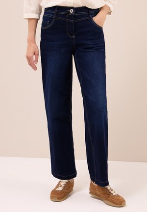Persoon in donkerblauwe straight-leg spijkerbroek, wit shirt met opgerolde mouwen en bruine veterschoenen, staand met één hand in de zak.