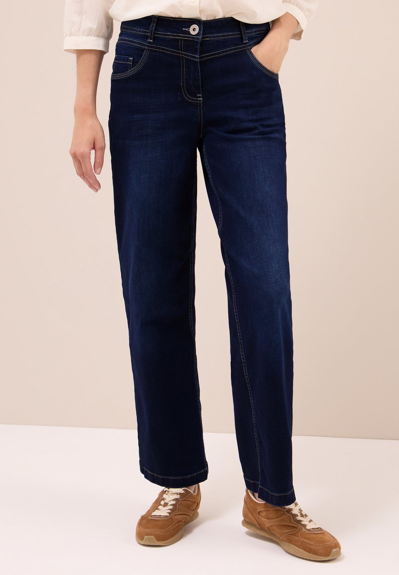 Persona vestida con jeans de pierna recta de color azul oscuro, camisa blanca con las mangas dobladas y zapatillas marrones con cordones, de pie con una mano en el bolsillo.