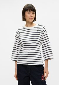 Ριγέ πουκάμισο σε λευκό και ναυτικό μπλε, με χαλαρή εφαρμογή και τρία τέταρτα μανίκια. Διαθέτει γραφικά σχέδια μπροστά και στρογγυλή λαιμόκοψη.