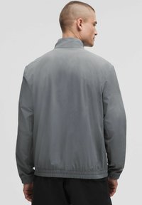 Veste légère grise avec un col haut, une taille élastique et des poignets élastiques. Présente une texture lisse et mate ainsi qu'un design minimaliste.