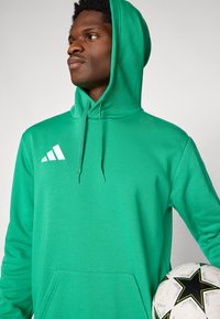 Πράσινο φούτερ με κουκούλα και εμπρόσθια τσέπη, με λευκό λογότυπο Adidas. Μια ασπρόμαυρη ποδοσφαιρική μπάλα κρατιέται στο ένα χέρι.