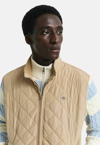 Gepatchte beigefarbene Weste mit hohem Kragen, ausgestattet mit einem Reißverschluss vorne, kombiniert mit einem rippenstrukturierten, gestreiften Pullover in Creme und Pastellblau.