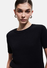 Sort strikket trøje med ribbet rund halsudskæring og korte ærmer. Stoffet har et tekstureret, tætsiddende væv. Tilbehøret inkluderer sølv hoops.