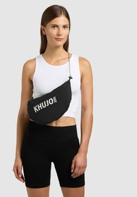 Kvinde iført hvid ærmeløs cropped top og sorte cykelshorts, bærende en sort crossbody taske med teksten "KHUJO 2001".