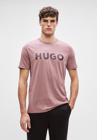 Ružové tričko vyrobené z bavlny, s okrúhlym výstrihom a krátkymi rukávmi. Na prednej strane sa nachádza výrazné čierne logo "HUGO". Nosené s tmavými nohavicami.