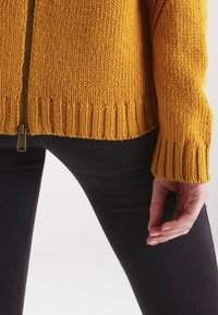 Gelbes Strickcardigan mit geripptem Saum und Bündchen, ausgestattet mit einem Reißverschluss. Texturiertes Material mit lockerer, bequemer Passform.