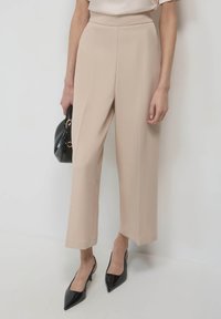 Pantalons beige à jambes larges avec une taille haute et une texture lisse. Associés à des escarpins noirs à bout pointu et à un petit sac à main noir.