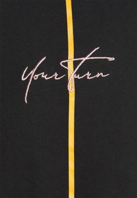 Zwarte stoffen shirt met een verticale gele streep. Aan de voorkant staat "Your Turn" geborduurd in roze cursief.