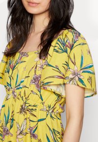 Vestido floral amarelo com mangas curtas, apresentando um decote quadrado e uma cintura franzida. Inclui padrões botânicos em rosa e verde.