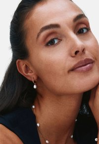 Pendiente de aro dorado con gota de perla blanca; la modelo tiene piel tersa y cabello oscuro, lleva un top negro y se ve un collar de perlas.