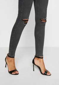 Svarta lackläder sandaler med hög klack, öppen tå, ankelrem och klackhöjd cirka 10 cm. Bärs med slitna gråa tajta jeans.