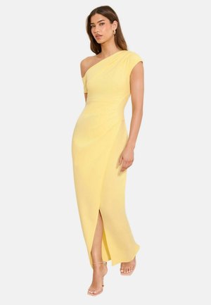 Kvinde iført en gul one-shoulder kjole med frontslids og beige strop-sandaler med høj hæl, stående foran en hvid baggrund.