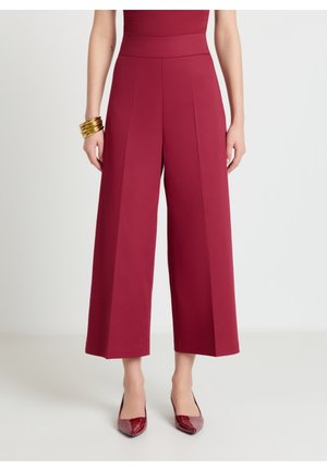 Donna che indossa pantaloni bordeaux a gamba larga e vita alta, tacchi appuntiti abbinati e braccialetti dorati al polso sinistro su sfondo bianco.