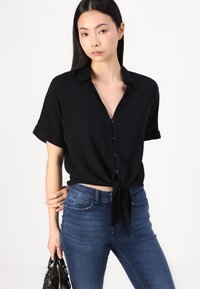 Black short-sleeve shirt with a collared neckline, button closure, and knotted hem. Paired with classic blue jeans and a black handbag.