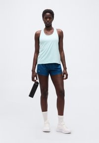 Femme athlétique debout, portant un débardeur bleu clair, un short bleu, des chaussettes blanches et des baskets, tenant une bouteille d'eau noire dans sa main droite.