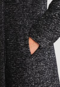 Närbild av en mörk, strukturerad kappa med en hand i fickan, som visar tygets detaljer och plaggets design.