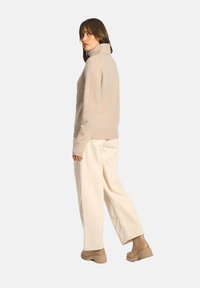 Beige Rollkragenpullover aus weichem, strukturiertem Stoff, kombiniert mit locker sitzenden, cremefarbenen Hosen und hellbraunen Knöchelstiefeln, von hinten betrachtet.