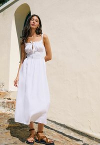 Vit maxiklänning med en åtsittande bustier, justerbara axelremmar och rynkad midja. Kombineras med bruna sandaletter med remmar på en texturerad yta.