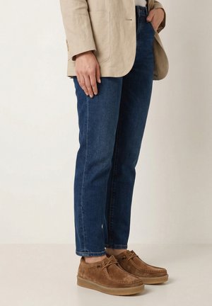 Person trägt blaue Jeans, beige Wildleder-Schnürschuhe und ein beiges Sakko und steht mit einer Hand in der Tasche vor einem schlichten Hintergrund.
