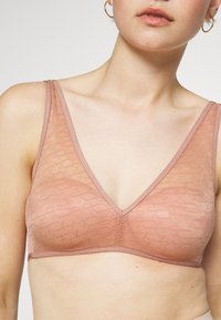 Hellrosa Bralette mit V-Ausschnitt-Design, aus transparenter Stoff gefertigt und mit einem subtilen strukturierten Muster sowie verstellbaren Trägern.