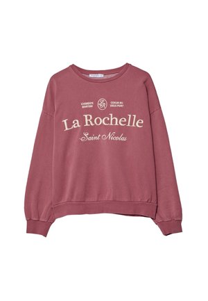 Person mit Händen in den Taschen, die einen mauvefarbenen Sweatshirt mit der Aufschrift „La Rochelle“, dunkle weite Hosen und weiße Turnschuhe trägt, steht drinnen.