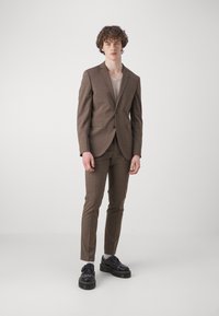 Costum cu carouri maro, cu o jachetă ajustată și pantaloni slim, asortat cu un pulover bej deschis și pantofi negri cu talpă groasă.