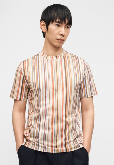 Νεαρός άνδρας που φοράει κοντομάνικη, πολύχρωμη μπλούζα με κάθετες ρίγες, με τα χέρια στις τσέπες του σκούρου παντελονιού, κοιτάζοντας κάτω με σκέψη.