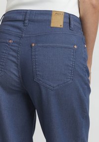 Vaqueros de mezclilla azul marino con una etiqueta de cuero etiquetada "PULZ", con acentos de botones en tono cobre y un diseño clásico de bolsillo trasero.