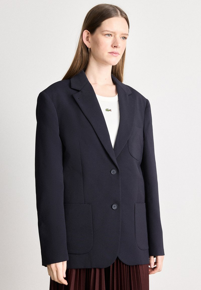 Blazer marine en tissu lisse, avec un col classique, deux poches avant et deux boutons sur le devant. Design simple et structuré.