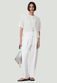 Top cropped de linho branco com mangas curtas, combinado com calças brancas largas. A modelo segura uma bolsa prateada e usa sandálias prateadas.