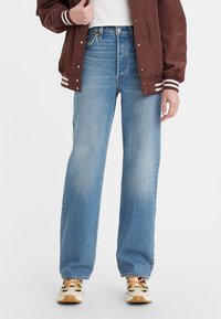 Un jean en denim bleu clair présente un design à jambe droite, un léger délavage et un style classique à cinq poches. Associé à une veste bomber en cuir marron et des baskets à motifs.