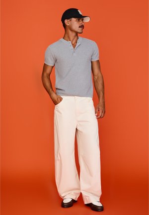 Mies, jolla on harmaa lyhythihainen henley, leveälahkeiset vaaleanbeiget housut, mustavalkoiset kengät ja musta lippalakki, seisoo oranssin taustan edessä.