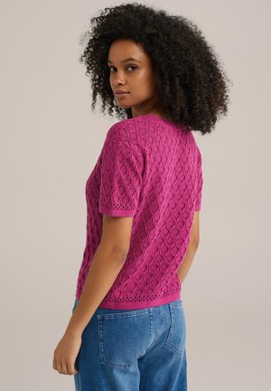 Top corto a maniche corte di colore fucsia, lavorato a maglia, caratterizzato da un motivo a diamante testurizzato e bordi a costine, indossato con jeans azzurri chiari.