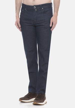 Uomo con jeans blu scuro a gamba dritta e sneakers marroni, in piedi davanti a uno sfondo chiaro e neutro.