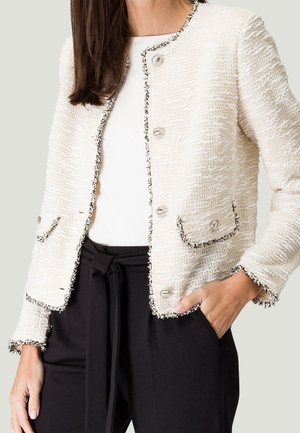 Frau trägt eine strukturierte cremefarbene Jacke mit schwarzem Besatz und Knöpfen, ein weißes Top und schwarze hochgeschnittene Hosen mit gebundenem Gürtel.