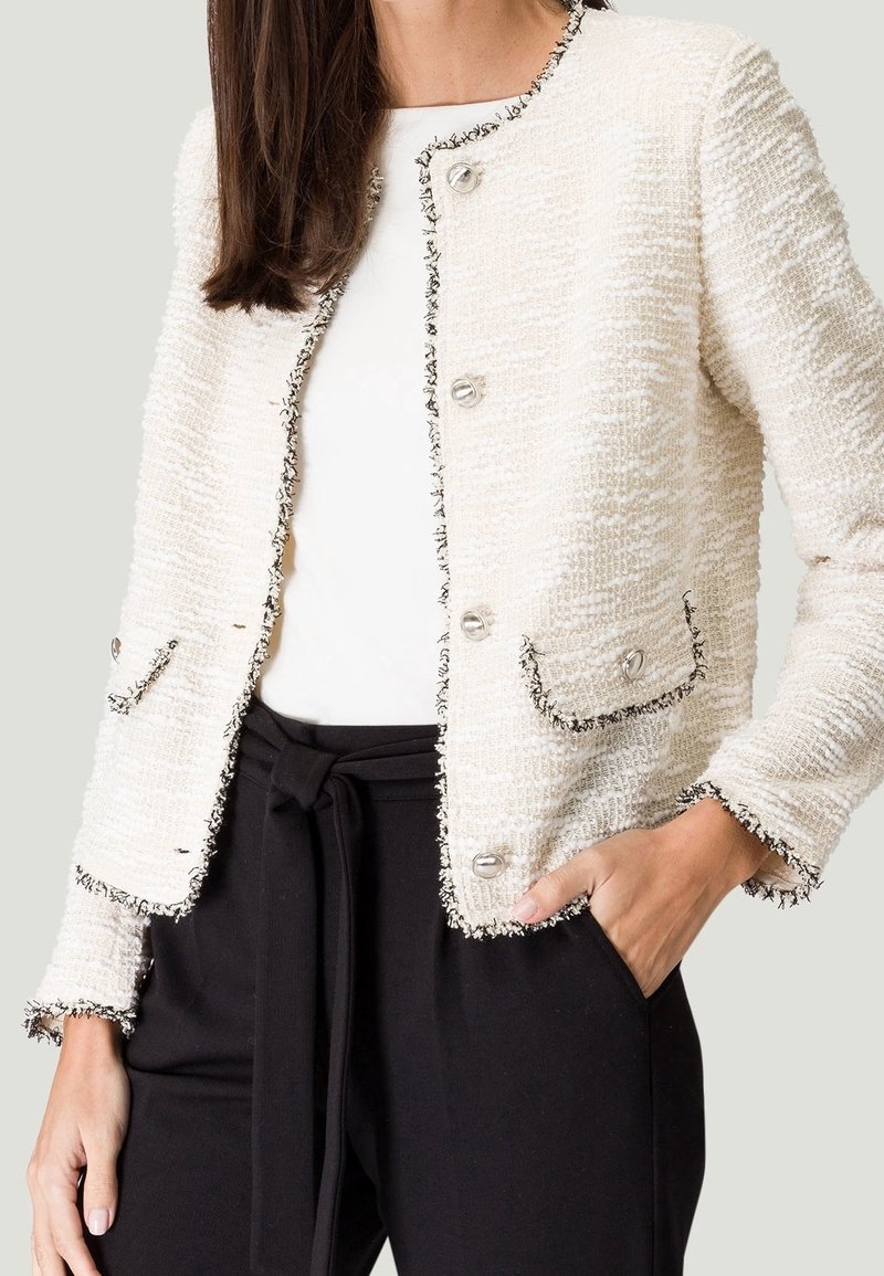 Femme portant une veste crème texturée avec des bordures et des boutons noirs, un haut blanc, et un pantalon noir taille haute avec une ceinture nouée.