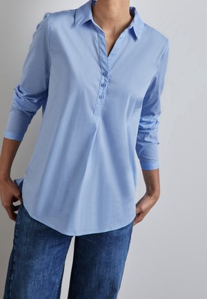 Chemise bleu clair à manches longues avec un col et un ourlet arrondi, fabriquée dans un tissu lisse.
