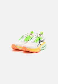 Chaussures de sport blanches, avec des lacets et des accents verts. Elles présentent une semelle orange vif et jaune, une tige en mesh et un logo noir sur le côté.