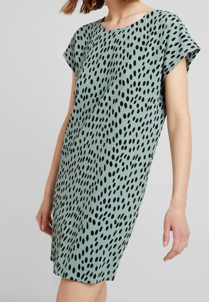 Femme portant une robe courte à manches courtes, de longueur genou, vert clair avec un motif de points noirs irréguliers et un col rond, devant un fond uni.